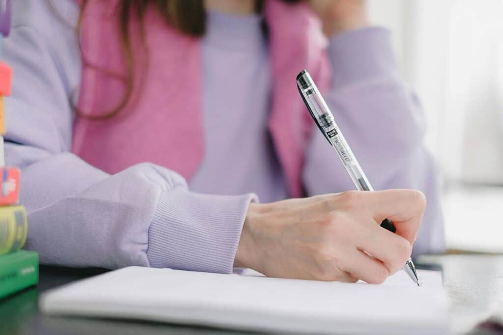 Woman in purple sweatshirt with a focus on her hand with a black gel pen writing in a planner 